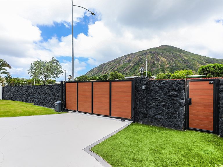The street-side gate at 654 Lunalilo Home Rd, a slatted wood gate set between stacked lava-rock walls, with Koko Head crater rising behind it under tradewind clouds