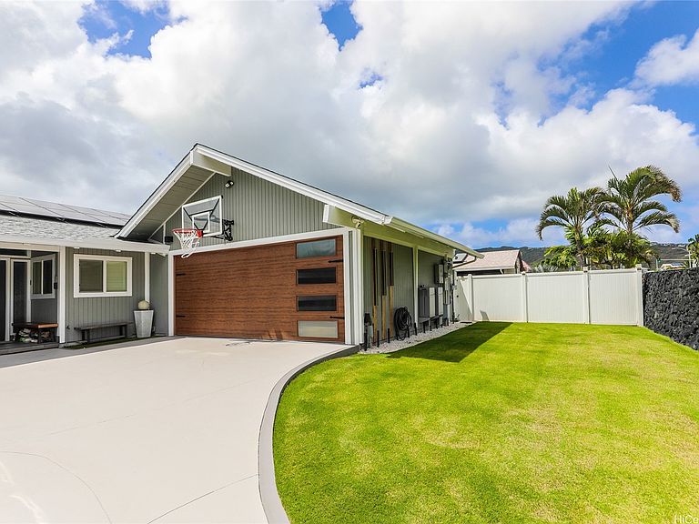 Three-quarter view of 654 Lunalilo Home Rd from the driveway, showing a wood garage door, a basketball hoop above it, palm trees and a glimpse of the ridge beyond