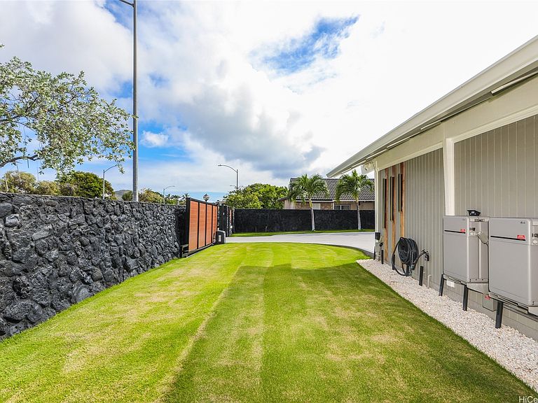 The side yard at 654 Lunalilo Home Rd: a long strip of bright green lawn between the house and a tall lava-rock wall, with tradewind clouds overhead